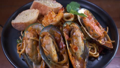 Köstliche-Muscheln-Mit-Meeresfrüchten-Mit-Spaghetti-Und-Knoblauchbrot,-Die-Sich-Auf-Einem-Schwarzen-Teller-Drehen
