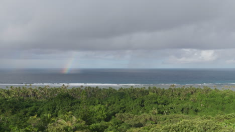 Panning-shot-above-tropical-coast-with-rainbow-falling-into-the-sea-off-Maré-Island,-New-Caledonia