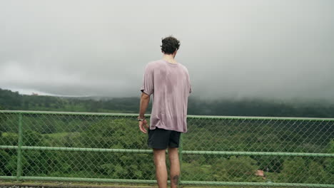 Man-stares-over-railing-at-overlook-as-shirt-blows-in-wind,-slow-motion