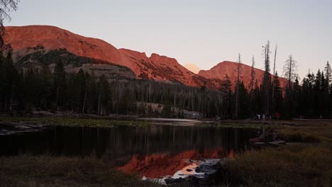 Landscape-tilting-up-shot-of-the-beautiful-Butterfly-Lake-surrounded-by-large-rocky-mountains-and-pine-trees-inside-of-the-Uinta-Wasatch-Cache-National-Forest-in-Utah-during-a-warm-summer-sunset
