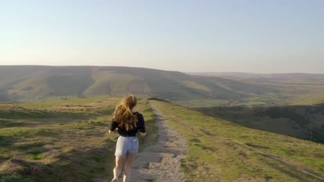 Stabilised-shot-of-young-blonde-woman-jogging-along-the-path-on-top-of-Mam-Tor,-Castleton,-Peak-District,-England,-view-of-green-rolling-hills-and-blue-skies