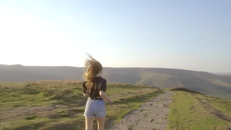 Stabilised-shot-of-young-blonde-woman-jogging-along-the-path-on-top-of-Mam-Tor,-Castleton,-Peak-District,-England-with-view-of-green-rolling-hills-and-blue-skies