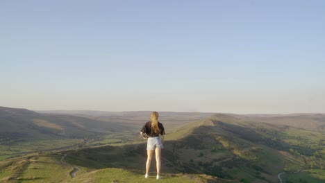Stabilised-shot-of-young-blonde-woman-jogging-along-the-path-on-top-of-Mam-Tor,-Castleton,-Peak-District,-England-before-stopping-to-admire-the-view-of-green-rolling-hills-and-blue-skies