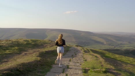 Stabilised-shot-of-young-blonde-woman-jogging-along-the-path-on-top-of-Mam-Tor,-Castleton,-Peak-District,-England