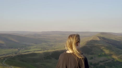 Stabilised-shot-of-young-blonde-woman-with-hair-over-one-shoulder-on-top-of-Mam-Tor,-Castleton,-Peak-District,-England-admiring-the-view-of-green-rolling-hills-and-blue-skies