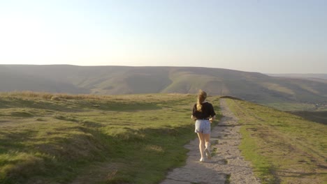 Stabilised-shot-of-young-blonde-woman-jogging-along-the-path-on-top-of-Mam-Tor,-Castleton,-Peak-District,-England