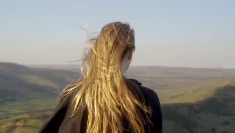 Stabilised-medium-shot-of-young-blonde-woman-standing-in-wind-on-top-of-Mam-Tor,-Castleton,-Peak-District,-England-before-stopping-to-admire-the-view-of-green-rolling-hills-and-blue-skies