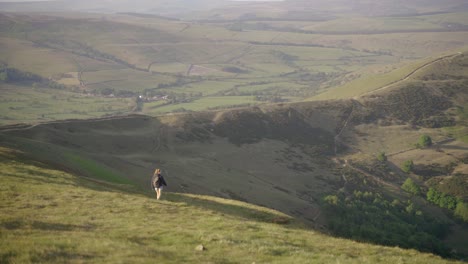 Handheld-sideways-dolly-of-young-blonde-woman-with-backpack-walking-down-the-side-of-Mam-Tor,-Castleton,-Peak-District,-England-at-sunset