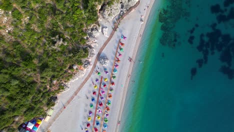 Colorful-umbrellas-on-beach-washed-by-turquoise-seawater-on-rocky-shore-of-Mediterranean-in-Albania