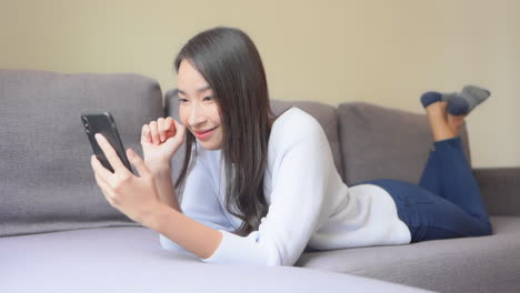 Junge-Glückliche-Asiatin,-Die-Auf-Der-Couch-Liegt-Und-Smartphone,-Soziales-Netzwerk-Oder-Selfie-Bildkonzept-Betrachtet,-Vollbild-Zeitlupe
