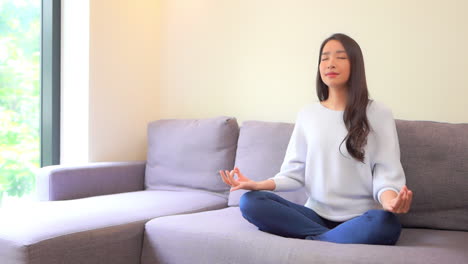 Asiatische-Junge-Frau,-Die-Auf-Ihrem-Sofa-Im-Wohnzimmer-Nahe-Einem-Fenster-Meditiert