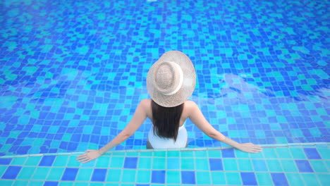 With-her-back-to-the-camera,-a-woman-in-a-white-one-piece-bathing-suit-and-straw-sunhat-leans-back-on-her-arms-as-she-surveys-the-pool