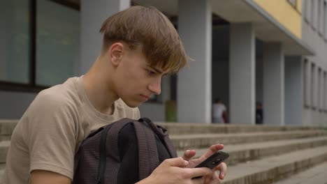 Youngster-sitting-in-front-of-the-school-and-scrolling-mobile-phone-with-his-backpack-in-front