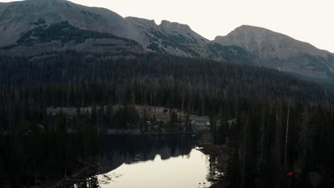 Aufsteigende-Luftdrohnenaufnahme-Des-Ruhigen-Schmetterlingssees-Mit-Seerosen-Im-Uinta-National-Forest-In-Utah-Mit-Großen-Felsigen-Bergen-Und-Pinien-Umgeben-An-Einem-Nebligen-Sommermorgen