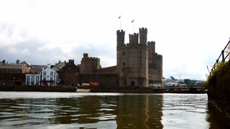Medieval-Caernarfon-castle-Welsh-tourist-town-stronghold-landmark-across-still-harbour-river-water