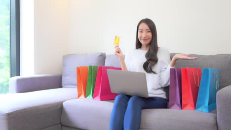 A-young-attractive-woman-with-a-laptop-on-her-knees-holds-a-golden-credit-card-up-in-one-hand-as-she-gestures-to-the-colorful-shopping-bags-that-surround-her-on-the-couch