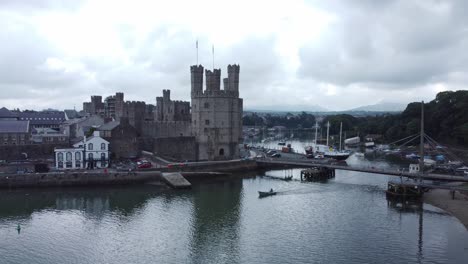 Ancient-Caernarfon-castle-Welsh-harbour-town-aerial-view-medieval-waterfront-landmark-descending-to-small-boat