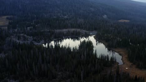 Aerial-drone-flying-over-the-tranquil-Butterfly-Lake-with-lily-pads-up-the-Uinta-National-Forest-in-Utah-with-large-Rocky-Mountains-and-pine-trees-surrounding-on-a-foggy-summer-morning-while-camping
