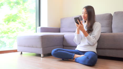 Young-Exotic-Woman-Swiping-Content-on-Smartphone-While-Sitting-on-the-Floor-of-Family-Home,-Smiling-and-Thinking,-Full-Frame-Slow-Motion
