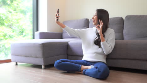 A-young-woman-sitting-cross-legged-on-the-floor-holds-her-smartphone-at-arm's-length-as-she-talks-and-waves