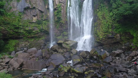 Aerial-of-Nauyaca-waterfalls-with-man-walking-at-base-in-rain-forest,-4K