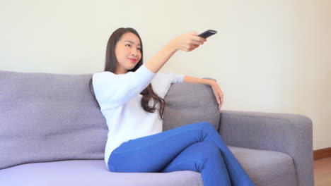 Pretty-asian-woman-sitting-in-sofa,-changing-television-channels-with-TV-remote,-full-frame-slow-motion