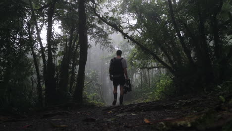 Slow-motion-shot-of-man-with-camera-gear-walking-down-foggy-rain-forest-path