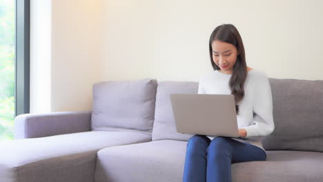Junge-Asiatische-Frau,-Die-Online-Von-Zu-Hause-Aus-Mit-Einem-Laptop-Im-Wohnzimmer-Arbeitet,-Vollbild-In-Zeitlupe