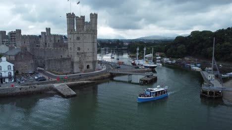 Sightseeing-boat-sailing-Caernarfon-castle-Welsh-harbour-town-river-aerial-view-descending-pull-back