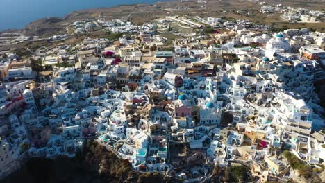 Luftflug-In-Richtung-Berühmtes-Griechisches-Dorf-Oia-Mit-Weißen-Höhlenhäusern-Und-Villen-In-Santorini,-Griechenland