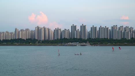 People-windsurfing-at-Han-River-on-Jamshil-skyscraper-apartment-cityscape-on-background-at-sunset,-Man-in-motorboat-taking-pictures-of-two-girls-on-paddleboards-in-the-middle-of-river-near-Ttukseom