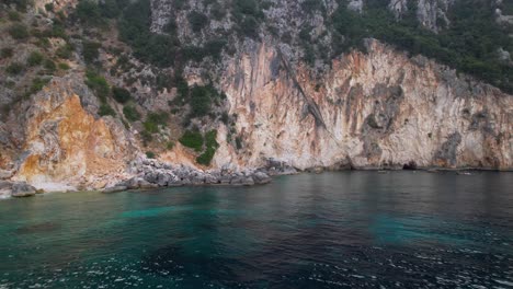 Rocky-slope-of-hills-and-natural-caves-on-shoreline-of-Mediterranean-in-Albania,-washed-by-emerald-sea-water