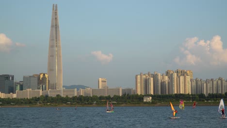 Windsurfistas-En-El-Río-Han,-Lotter-World-Tower-En-El-Fondo-Al-Atardecer,-Seúl,-Corea-Del-Sur
