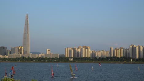 Group-of-Unrecognizable-Windsurfers-Surfing-on-Han-River,-Seoul-at-Sunset,-famous-Lotter-Tower-landmark-Building-on-Background