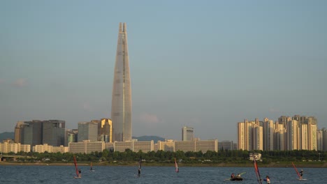 Group-of-People-windsurfing-on-Han-river-next-to-famous-korean-landmark-Lotte-World-Tower-at-golden-hour---copy-space---Jamsil,-Tukseom-Park-surf-club