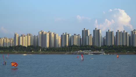 People-windsurfing-on-Han-river-at-sunset-on-complex-high-apartment-background,-Ttukseom-Surfing-club