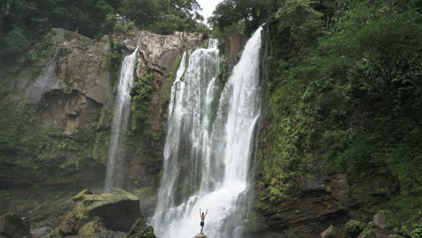 Man-stands-with-arms-raised-in-joy-in-front-of-large-rain-forest-waterfall