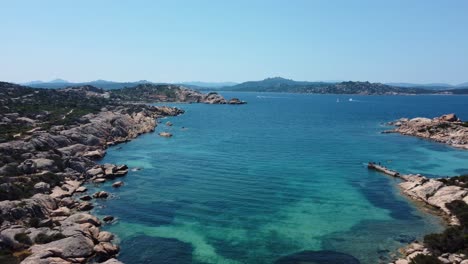 Flying-along-an-idyllic-natural-coast-beach-rock-bay-at-La-Maddalena-on-the-tourist-vacation-island-Sardinia-in-Italy-with-sun,-clear-blue-turquoise-and-calm-water