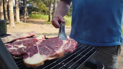Low-angle-view-of-man-placing-large-steaks-on-grill-while-camping