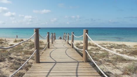 Male-tourist-with-Bermuda-shorts,-polo-shirt,-backpack-and-a-cap-walking-on-a-wooden-bridge-coming-from-a-beautiful-azure-beach