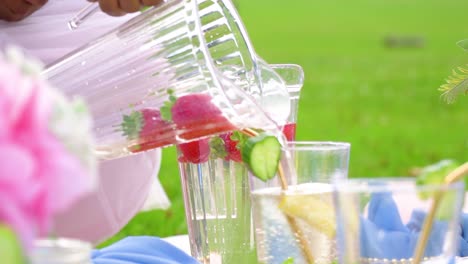 Plastikglas-Mit-Erdbeeren-Und-Wasser-Wird-Von-Einer-Braunen-Hautfrau-Für-Eine-Gesunde-Lebensstilparty-In-Der-Natur-Gegossen