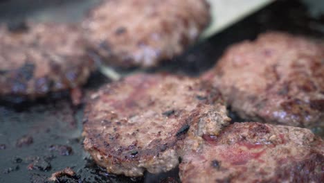 Hamburger-beef-meat-sausage-pork-patties-grilling-on-cast-iron-skillet-grill-cooked-with-propane-at-campsite-outdoor-barbecue-with-spatula-moving-around,-close-up-shot
