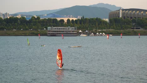 People-Windsurfing-on-Han-river-at-Sunset-near-Ttukseom-Park,-Jamsil-Sports-Complex-or-Jamsil-Arena-and-distant-mountains-on-background