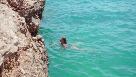 Mujer-Sexy-Nadando-En-Un-Mar-Azul-Brillante-Hacia-La-Costa-Escarpada-En-Verano