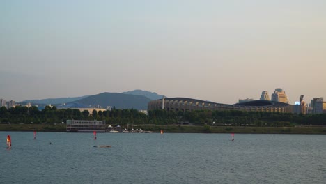 People-Windsurfing-on-Han-river-at-Sunset-near-Ttukseom-Park,-Jamsil-Sports-Complex-or-Jamsil-Arena-and-distant-mountains-on-background---wide-angle-shot-copy-space