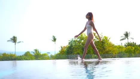 Slow-motion-view-of-attractive-Asian-model-in-swimsuit-walking-in-shallow-water-of-infinity-pool-at-tropical-luxury-resort-with-view-of-ocean-and-mountains-in-background