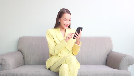 Attraktive-Asiatische-Geschäftsfrau-In-Hellgelber-Bürokleidung-Mit-Smartphone,-Während-Sie-Auf-Der-Couch-Sitzt