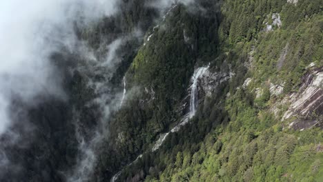 Vista-Aérea-De-Drones-De-Una-Cascada-Y-Un-Torrente-En-La-Ladera-De-Una-Montaña,-Una-Gran-Nube-Ocultando-El-Bosque-Debajo-En-La-Sombra-En-El-Norte-De-Italia