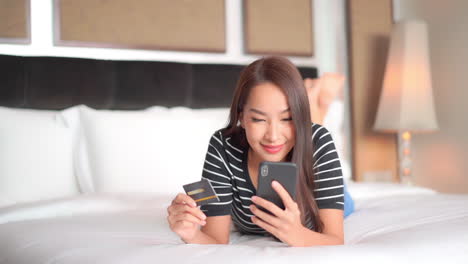 Weibliches-Asiatisches-Modell,-Das-Auf-Bett-Liegt-Und-Kredit--Oder-Debitkarte-Und-Smartphone-Lächelnd-Hält