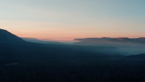Beautiful-rising-aerial-drone-shot-of-the-stunning-wild-Uinta-Wasatch-Cache-National-Forest-in-Utah-with-large-pine-trees-below-and-stunning-mountains-covered-in-mist-during-a-summer-sunrise